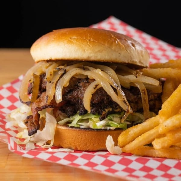 Matty G's Steakburger with caramelized onions, bacon, lettuce, and fries on red checkered paper.