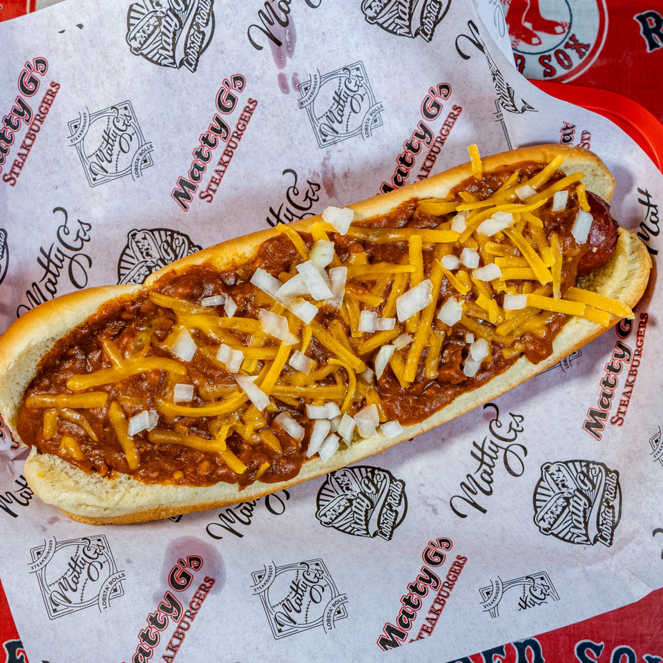 A Matty G's chili cheese dog with diced onions on branded paper in a red basket.