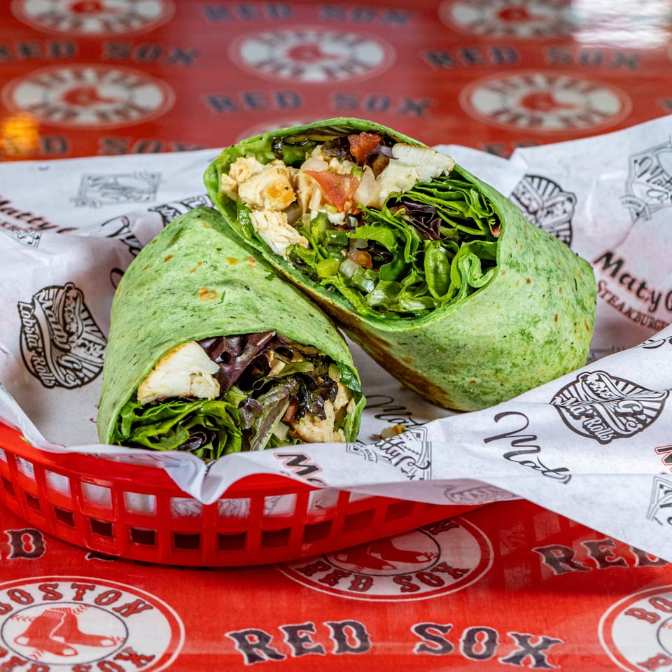 Two green wraps filled with chicken, lettuce, and tomatoes in a red basket on a Boston Red Sox tablecloth.