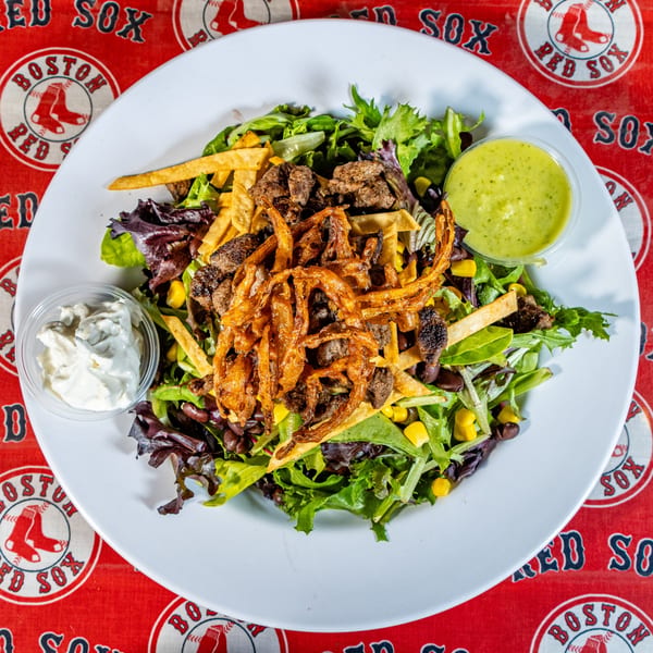 A Matty G's Steakburger salad with steak, fried onions, corn, and beans on a Boston Red Sox tablecloth.
