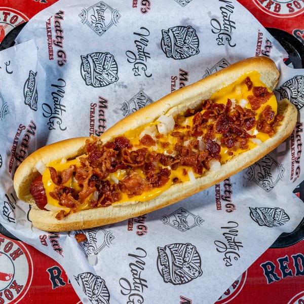 Matty G's hot dog with melted cheese, bacon bits, and diced onions on branded paper.