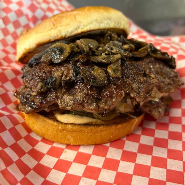 Matty G's mushroom steakburger with two patties, sauce, and pickles on a red and white checkered paper.