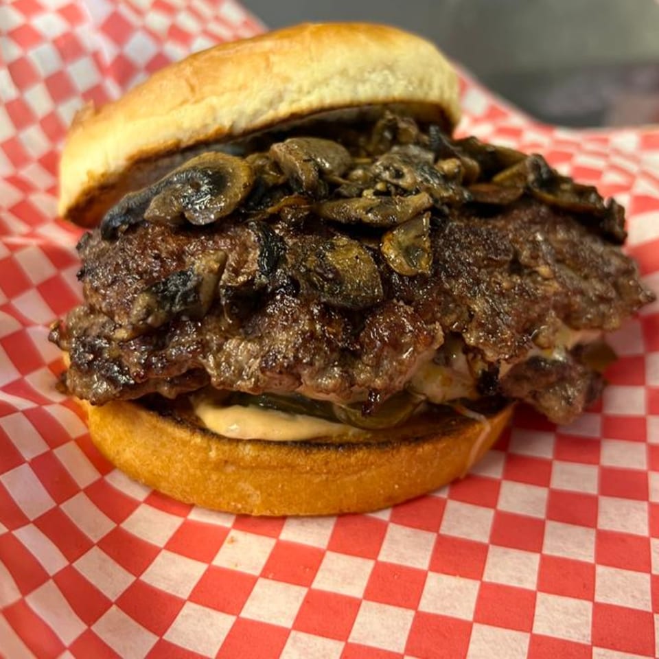 Matty G's mushroom steakburger with two patties, sauce, and pickles on a red and white checkered paper.