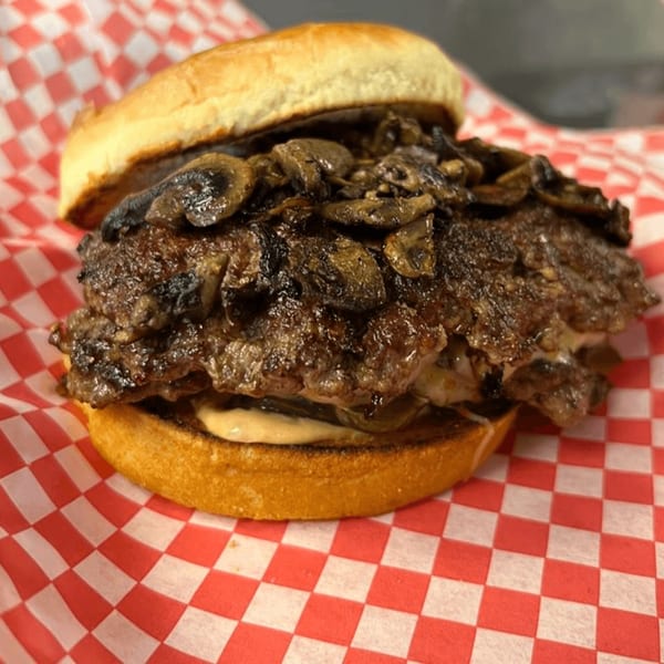 Matty G's Steakburger with grilled mushrooms and sauce on a toasted bun, on red and white checkered paper.