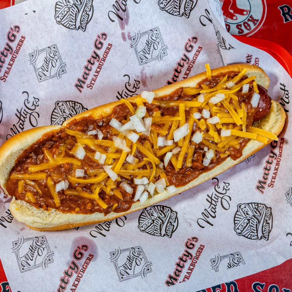 A Matty G's chili cheese hot dog with diced onions on branded paper.