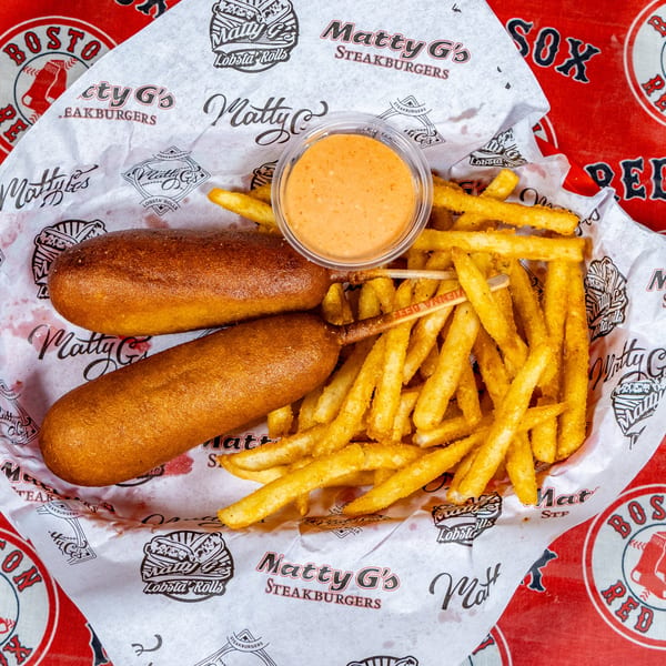 Two Matty G's corn dogs and a side of fries with dipping sauce in a branded paper-lined basket.