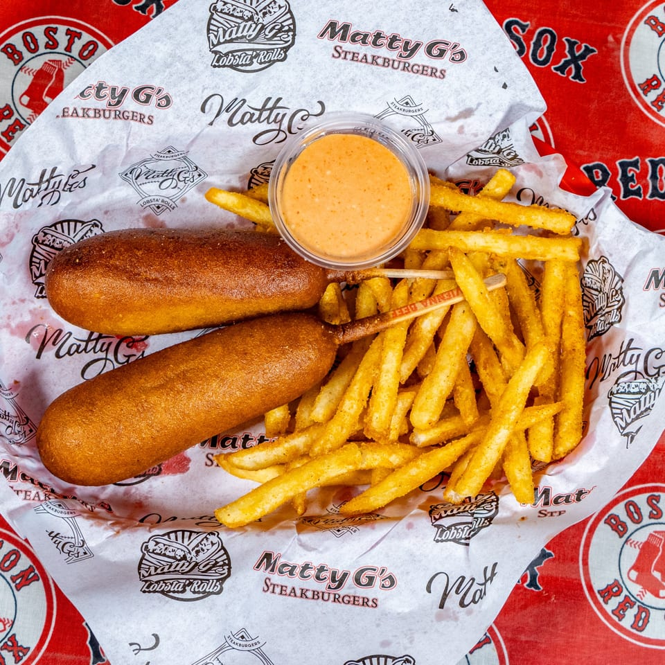 Two corn dogs, a side of Matty G's seasoned fries, and dipping sauce in a branded paper-lined basket.