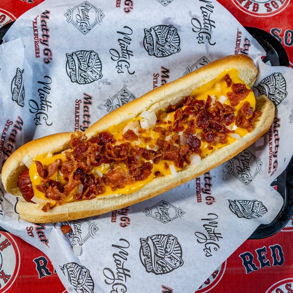 Matty G's hot dog with melted cheese, bacon bits, and diced onions on branded paper.