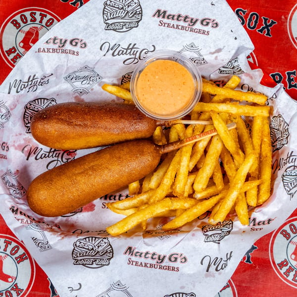 Two corn dogs, a side of fries, and dipping sauce in a basket with Matty G's branded paper.